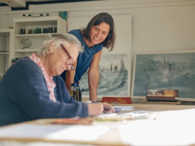 Ordenswerke_Suchthilfe_SozialeTeilhabe_Adelsdorf_LauferMuehle_Arbeitsangebote_4 Zwei Frauen in einem hellen Atelier. Eine ältere Frau sitzt am Tisch und zeichnet konzentriert, während eine jüngere Frau hinter ihr steht und freundlich lächelt. Im Hintergrund sind gerahmte Bilder an der Wand zu sehen.
