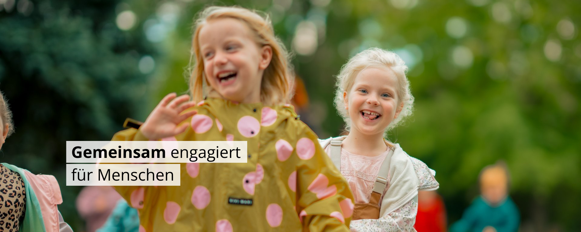 Zwei lachende Mädchen rennen spielerisch im Freien. Ein weißes Textfeld darüber lautet: „Gemeinsam engagiert für Menschen“. Im verschwommenen Hintergrund sind weitere spielende Kinder und grüne Bäume zu erkennen.