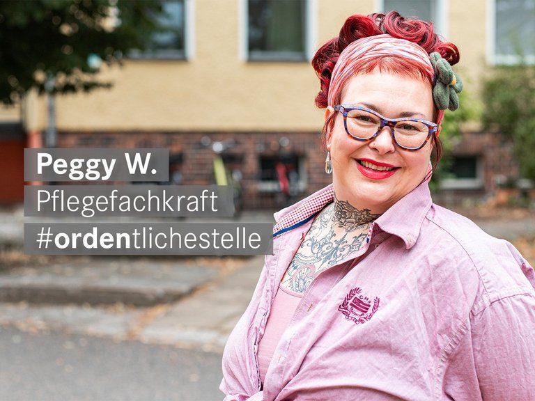 Pflegefachkraft mit roten Haaren Brille und Tattoos laechelt freundlich in die Kamera