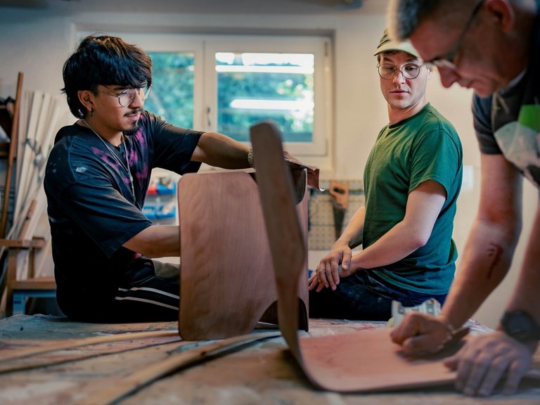 Drei Männer arbeiten in einer Werkstatt an einem großen Holzteil. Zwei Männer halten und bearbeiten das Holz, während der dritte Mann aufmerksam zuschaut.
