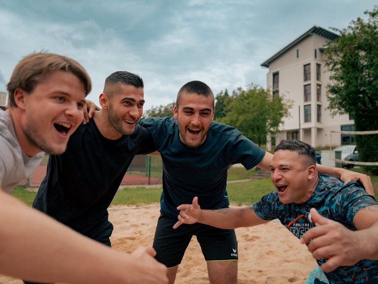 Vier fröhliche junge Männer stehen eng beieinander auf einem Beachvolleyballplatz und lachen in die Kamera, während sie sich für ein Gruppenfoto die Arme um die Schultern legen.