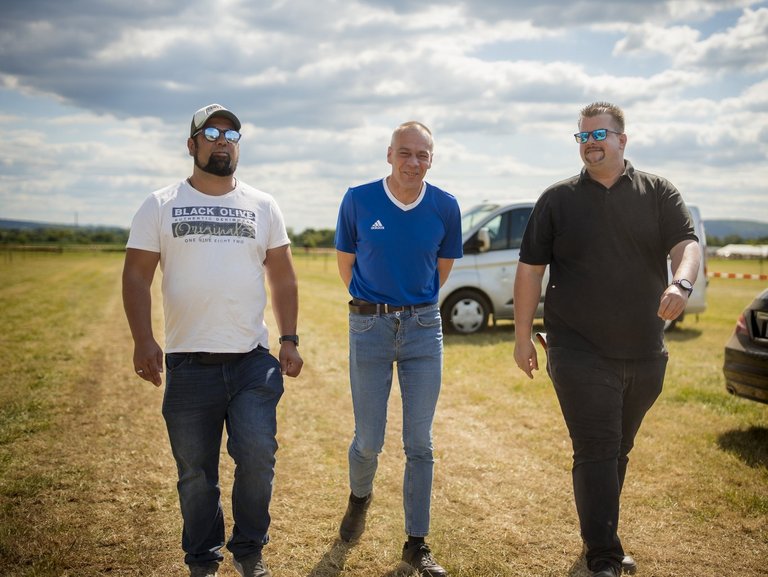 Drei Männer gehen nebeneinander über ein grasbewachsenes Feld. Im Hintergrund sind ein weißer Van und ein schwarzes Auto geparkt. Der Himmel ist bewölkt.