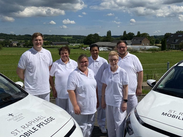 Das Team des Ambulanten Pflegedienstes steht zwischen zwei Ambulanten Fahrzeugen im Freien. 